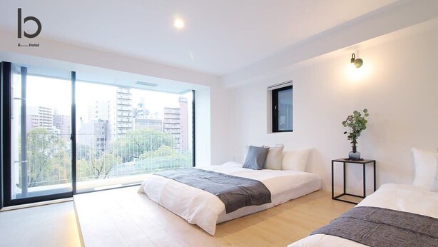 A modern hotel room with two beds, a large window with a city view, a small table with a plant, and a wall-mounted light fixture. - b hotel 401 Premium City View Apartment (Hiroshima City)