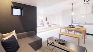 A modern hotel room with a grey sofa, a small table, a dining area with a wooden table and chairs, and a kitchen area with white cabinets and appliances. - b hotel 401 Premium City View Apartment (Hiroshima City)