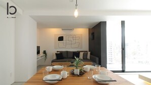 Uma área de jantar moderna com mesa de madeira posta para quatro pessoas, uma planta em vaso e vista para a cidade através de grandes janelas.