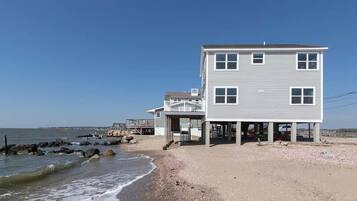 A two-story beach house with a balcony, situated on a rocky shoreline.