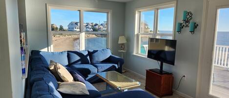 Una sala de estar con un sofá azul, un televisor y grandes ventanales con vistas a la playa.