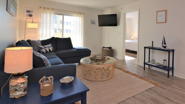 A living room with a sofa, a glass coffee table, a lamp, and a TV mounted on the wall.