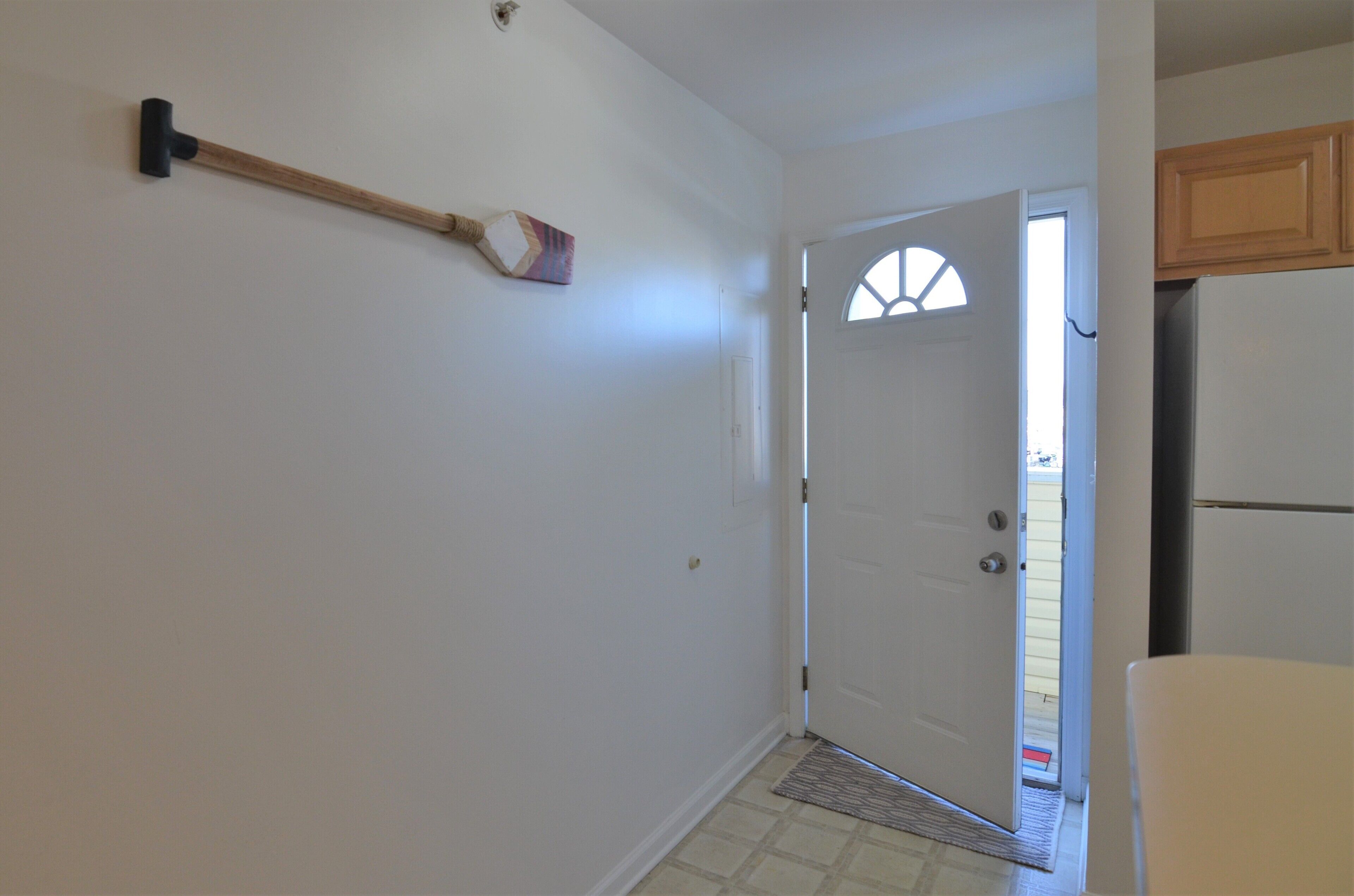 A hallway with a broom leaning against the wall, a door with a window, and a refrigerator in the adjacent room.