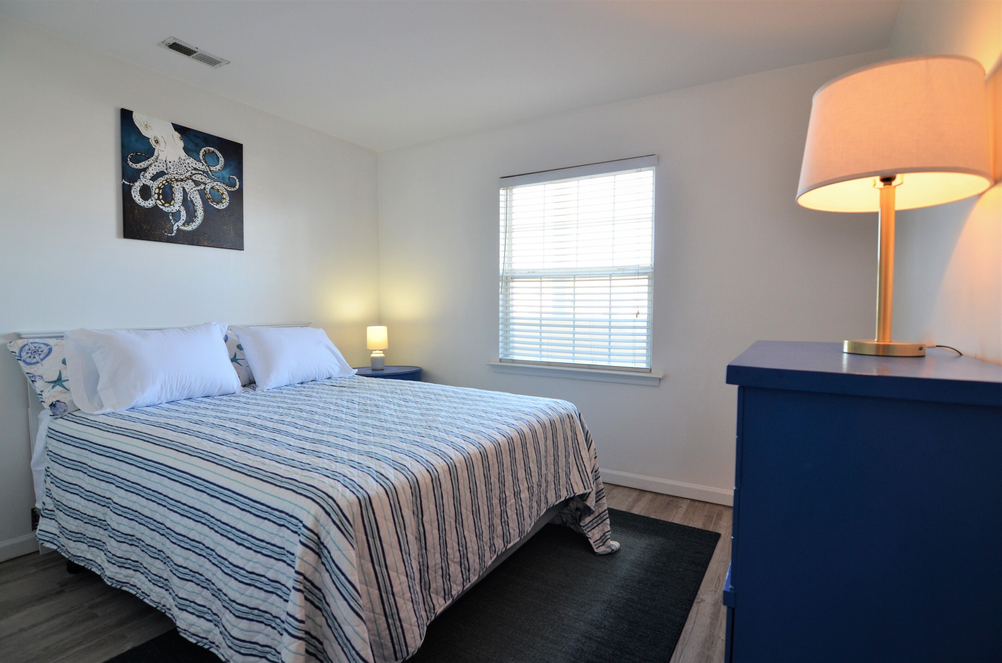 A bedroom with a bed, a nightstand, a lamp, a window, and a painting on the wall.