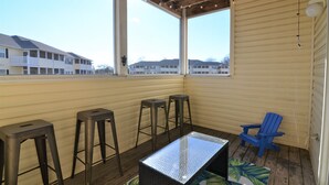 A covered outdoor seating area with a table and stools, a blue chair, and large windows overlooking a building.