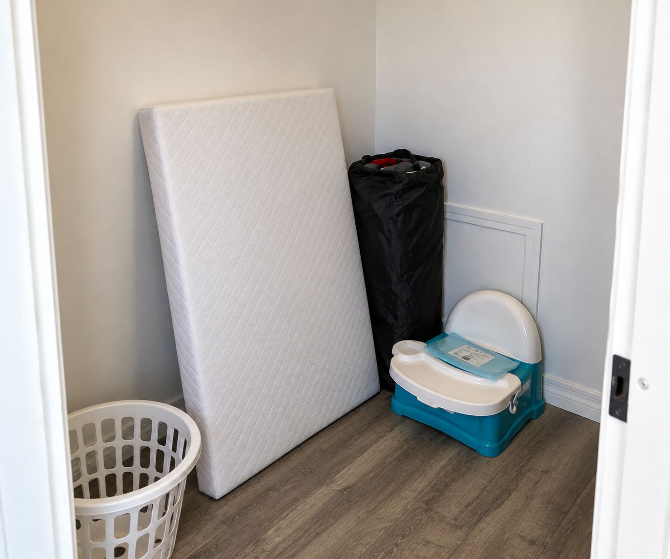 Een kleine badkamer met een witte matras, een draagbaar toilet, een prullenbak en een wasmand.