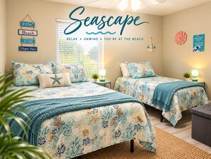 A bedroom with two beds, each with matching floral bedding and starfish pillows. There is a wall-mounted sign that reads 'Seascape' and a decorative anchor.