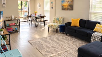 A living room with a dark sofa, a patterned rug, a dining area with chairs and a table, and a view of the outdoors through a sliding door.