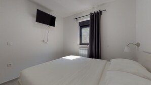 Ett sovrum med en säng, en väggmonterad TV, ett fönster med gardiner, ett element och en vägglampa.