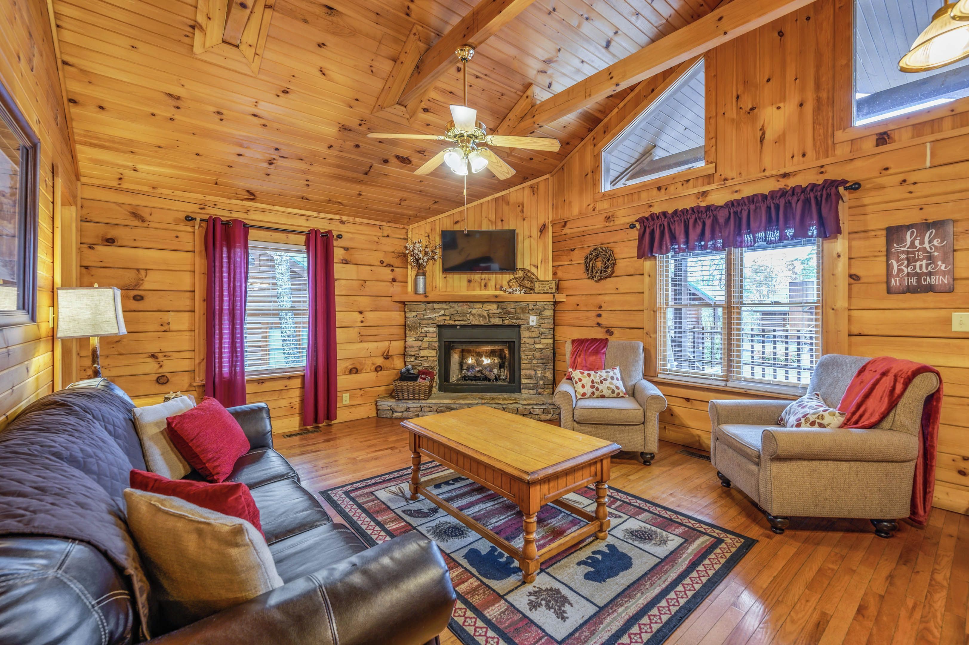 A cozy cabin interior with a stone fireplace, wooden walls, a ceiling fan, and a large window.