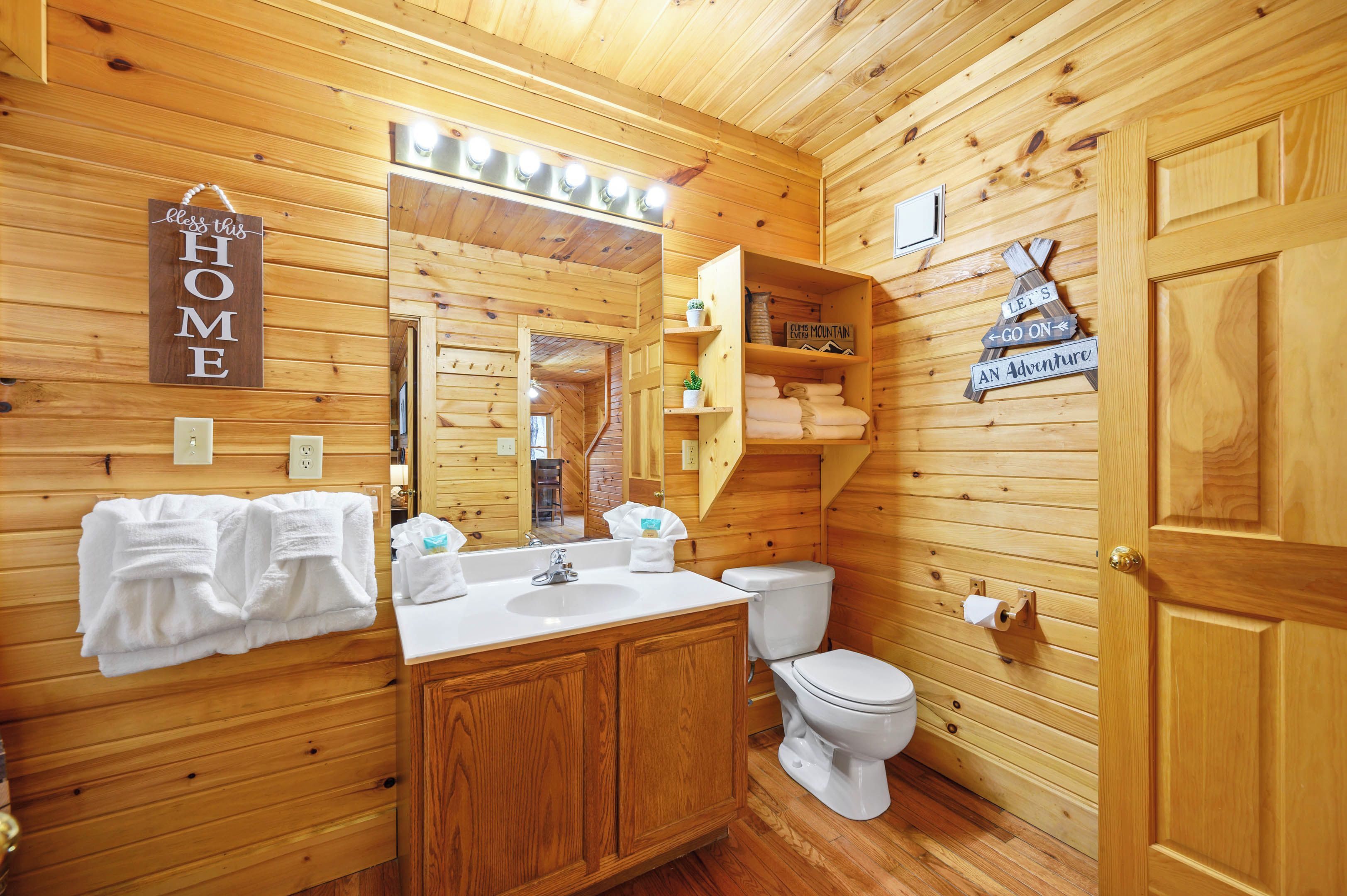 Une salle de bain rustique, avec des murs en bois, comprenant un meuble-lavabo en bois, des toilettes et un miroir. Des serviettes sont accrochées au mur, et une pancarte indique : « Partons à l’aventure ! »