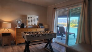 Una habitación con mesa de futbolín, escritorio con computadora, ventilador y vista a un cuerpo de agua a través de amplias ventanas.