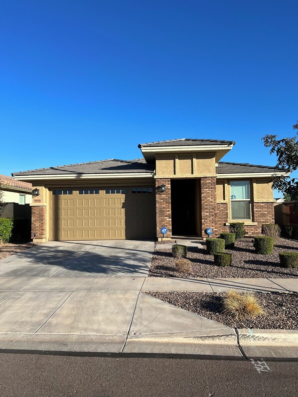 A two-story house with a beige garage door, a brick facade, and a small front yard with shrubs and gravel.