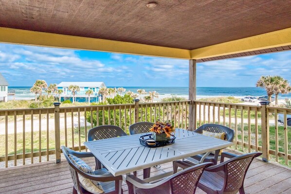 A covered outdoor area with a table and chairs, overlooking a beach with palm trees and a building in the background.