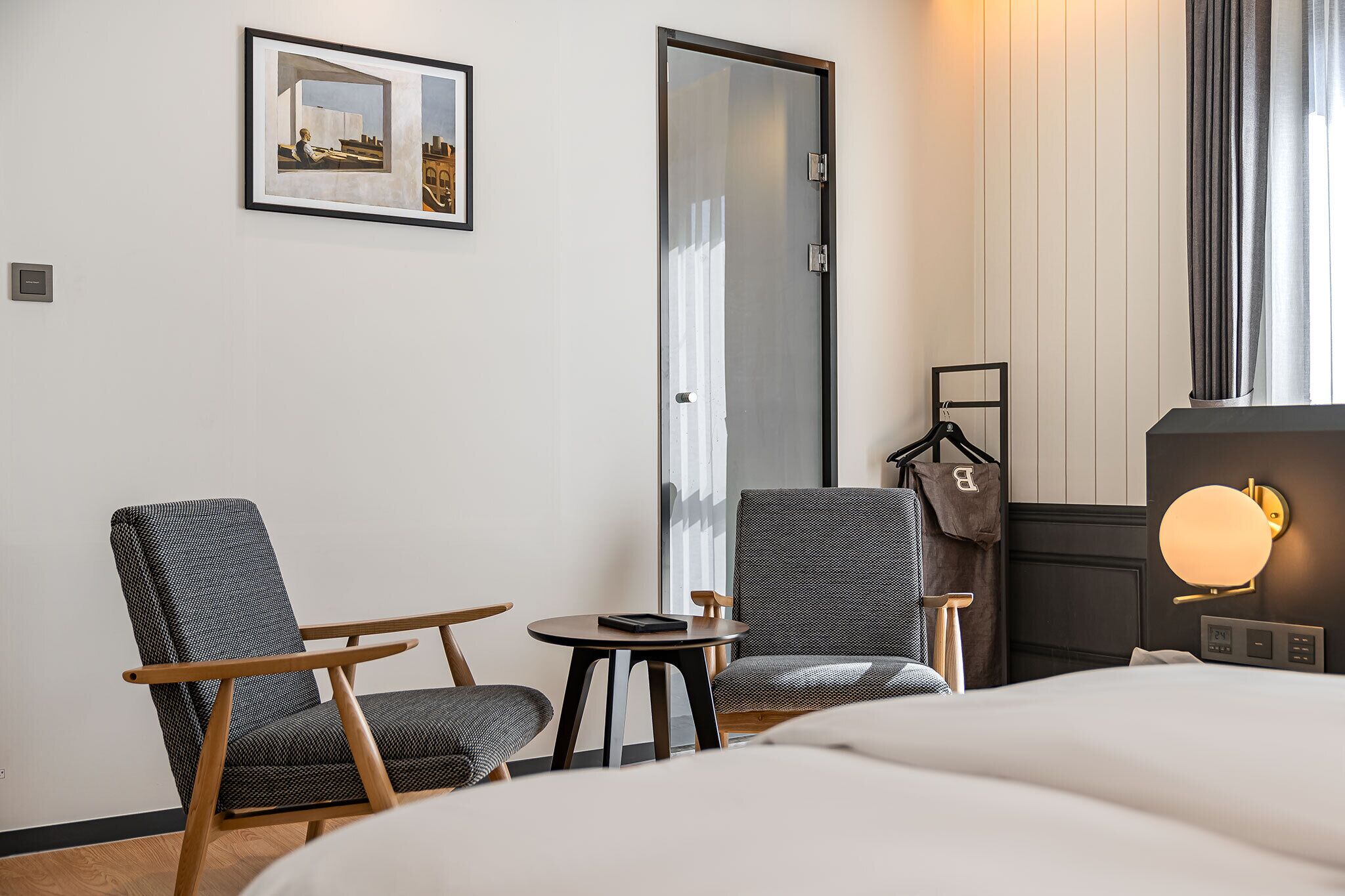 Habitación de hotel con una cama, dos sillones, una mesita, perchero y un cuadro en la pared.