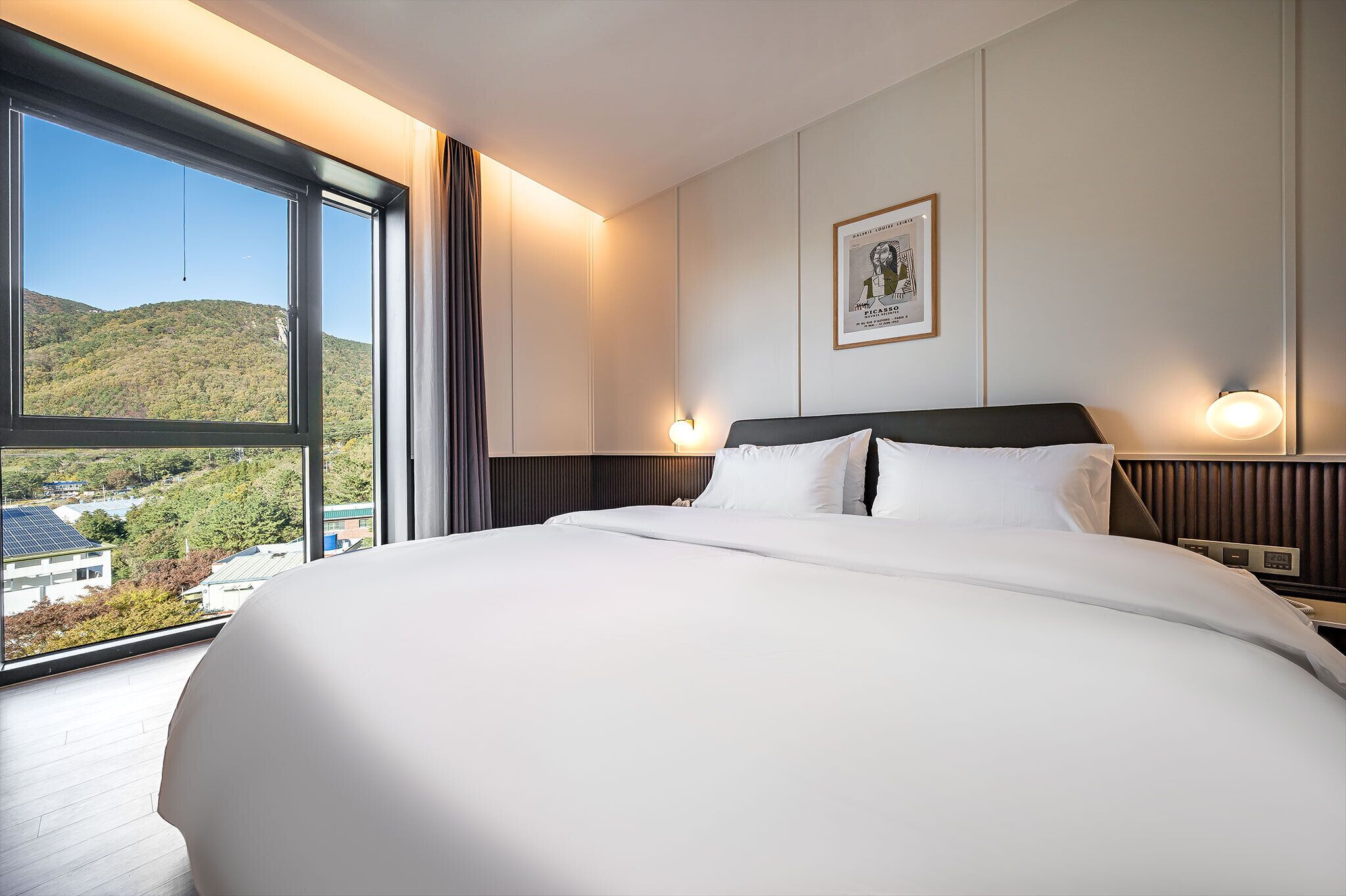 Une chambre d’hôtel avec un grand lit, deux lampes de chevet, un tableau encadré au mur et une vue sur un paysage verdoyant par une grande fenêtre.