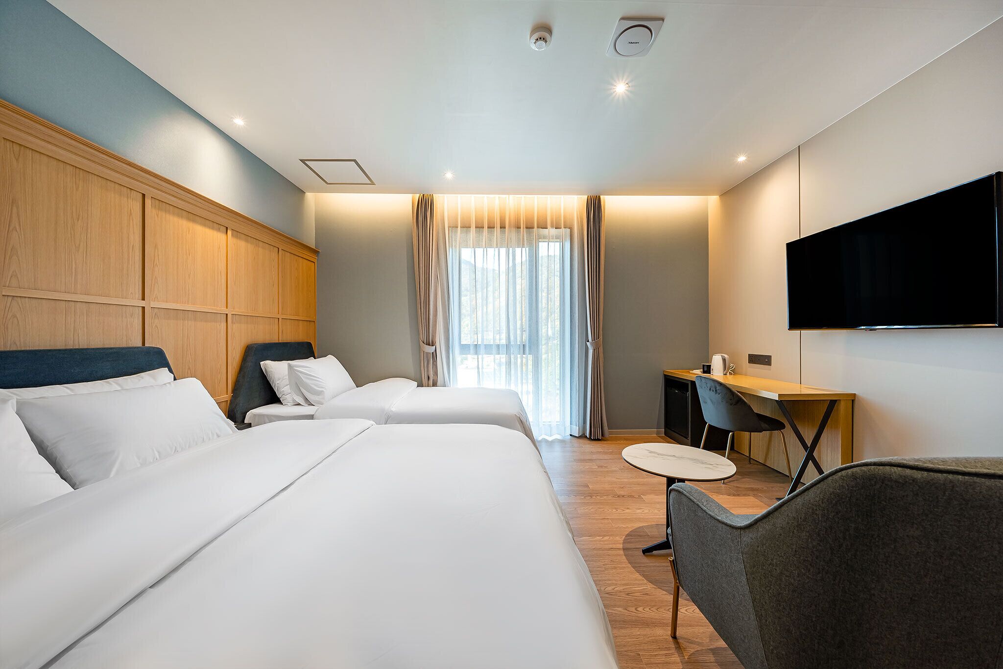 A hotel room with two beds, a desk, a chair, a flat-screen TV, and a window with curtains.