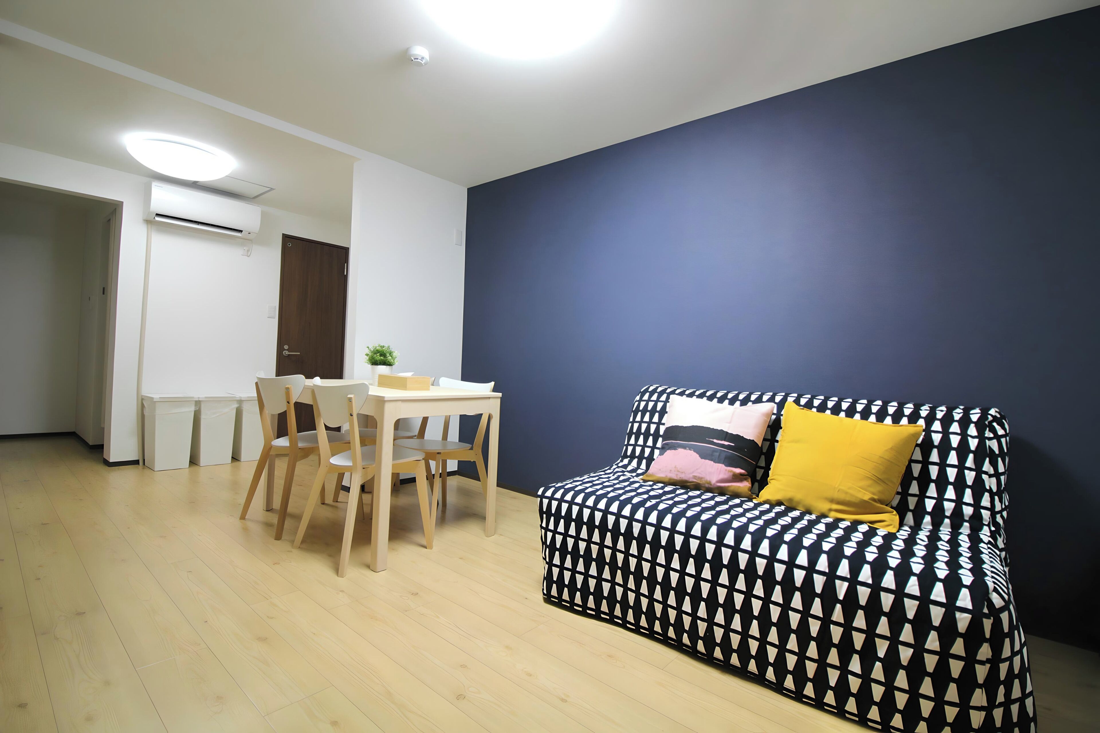A living room with a dark blue wall, a patterned sofa with a yellow pillow, a wooden dining table with chairs, and an air conditioning unit on the wall.