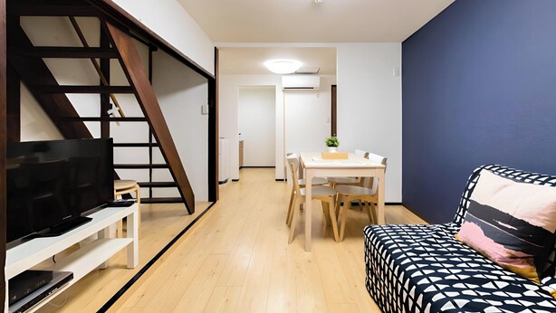 Uma sala de estar moderna com uma TV sobre um suporte branco, um sofá com estampa, uma mesa de jantar com cadeiras e uma escada.
