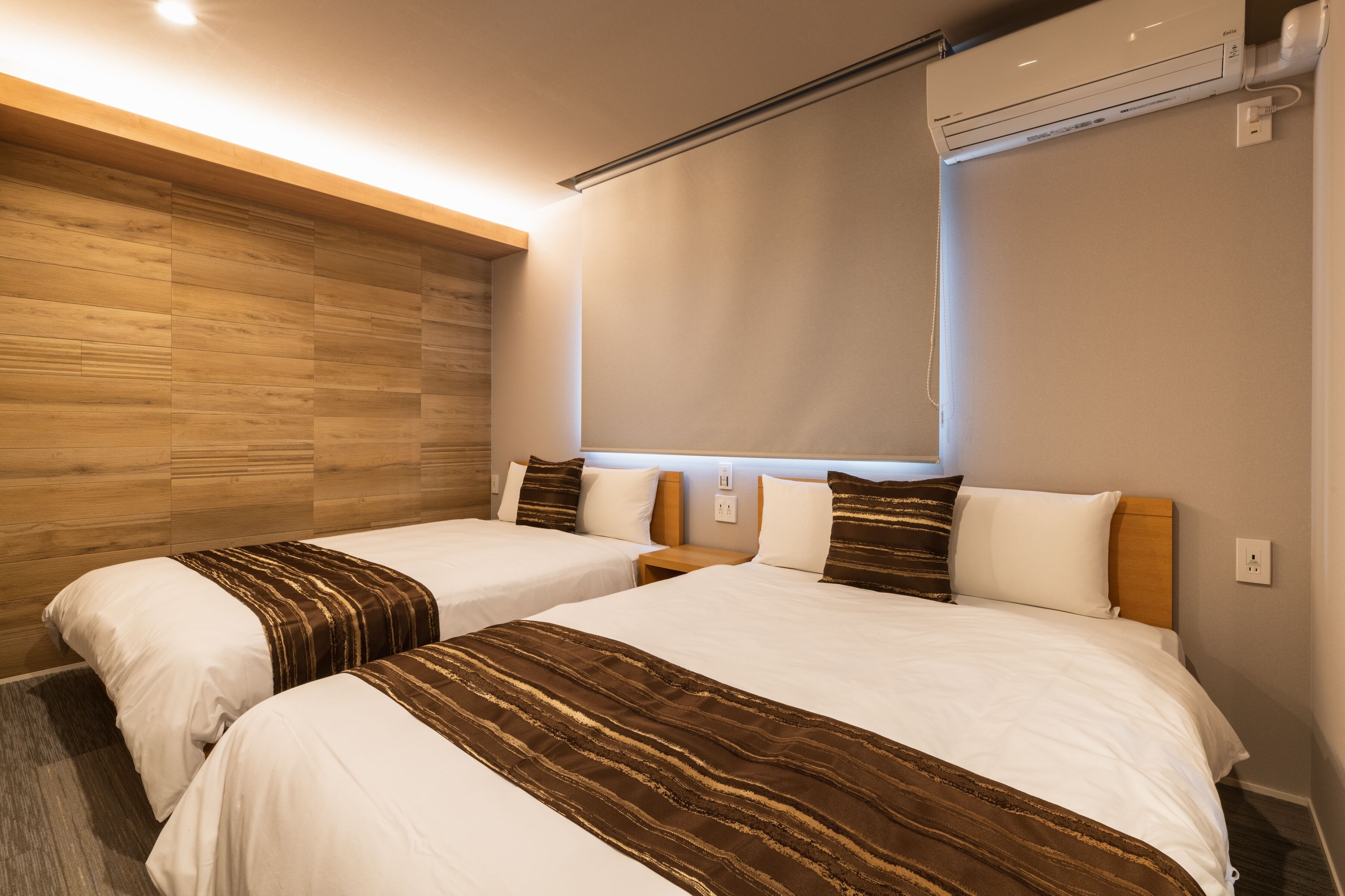 A hotel room with two beds, wooden paneling on the left wall, a ceiling light, and an air conditioning unit.