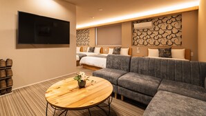 A modern hotel room with a large gray sofa, a round wooden coffee table, and a flat-screen TV mounted on the wall. - Grand Base Urakami (Nagasaki)