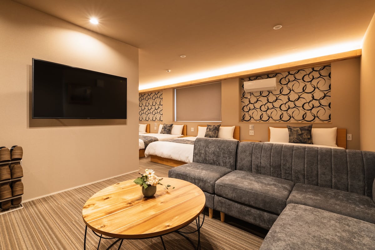 A modern hotel room with a large gray sofa, a round wooden coffee table, and a flat-screen TV mounted on the wall.