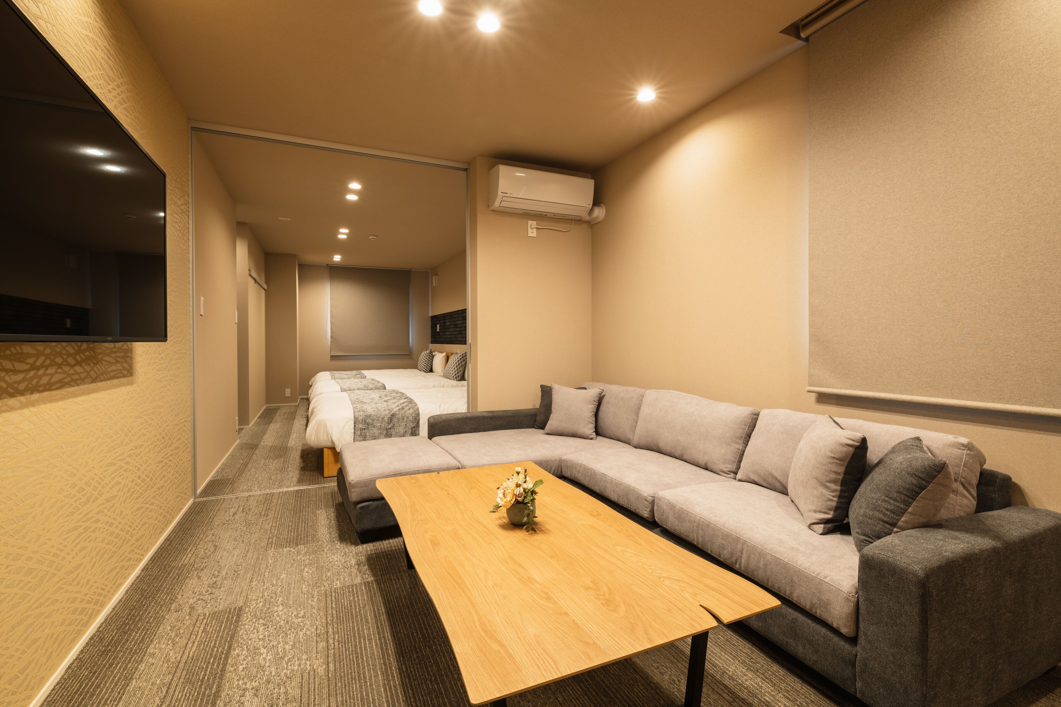 Habitación de hotel moderna con un sofá grande, una mesa de centro de madera y una cama al fondo.