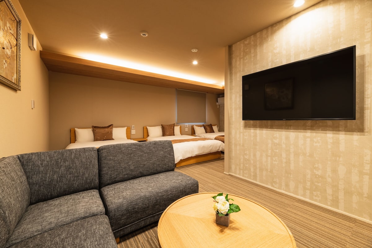 A hotel room with a sofa, a bed, a TV mounted on the wall, and a small table with a flower vase.