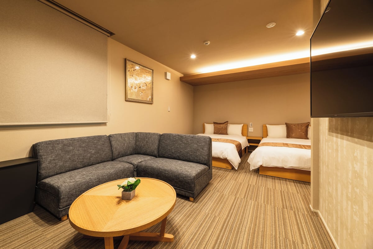 A hotel room with a gray sectional sofa, a round wooden table with a plant, a bed with white and brown bedding, and a flat-screen TV mounted on the wall.