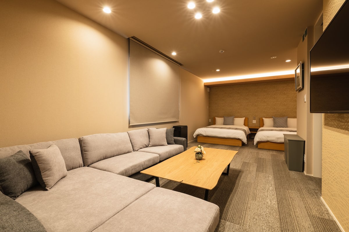 A modern hotel room with a large sofa, a wooden coffee table, a bed with two pillows, a flat-screen TV, and a small speaker.