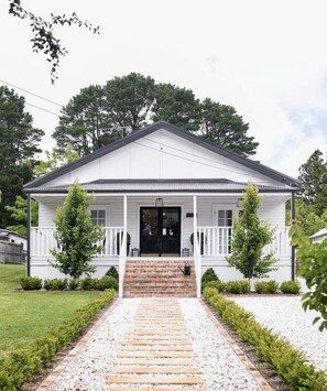 A white single-story house with a front porch, a black front door, and a small front yard with a gravel pathway leading to the door. - Luxury Hamptons Home (Leura)