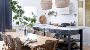 A modern kitchen with a central island, dining table, wicker chairs, and hanging rattan light fixtures.