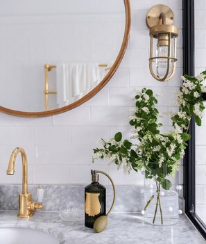 A bathroom with a gold faucet, a round mirror, a wall-mounted light fixture, and a vase with white flowers. - Luxury Hamptons Home (Leura)