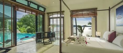 A spacious bedroom with a four-poster bed, a view of the ocean, and a pool area visible through large sliding doors.