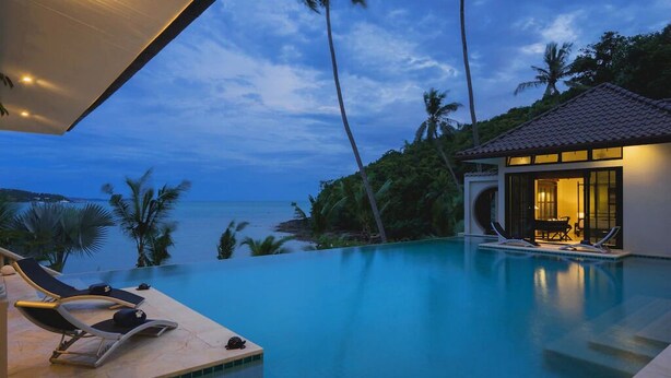 Una piscina infinita con sillones de descanso, un pequeño edificio con techo de tejas y vista al océano.