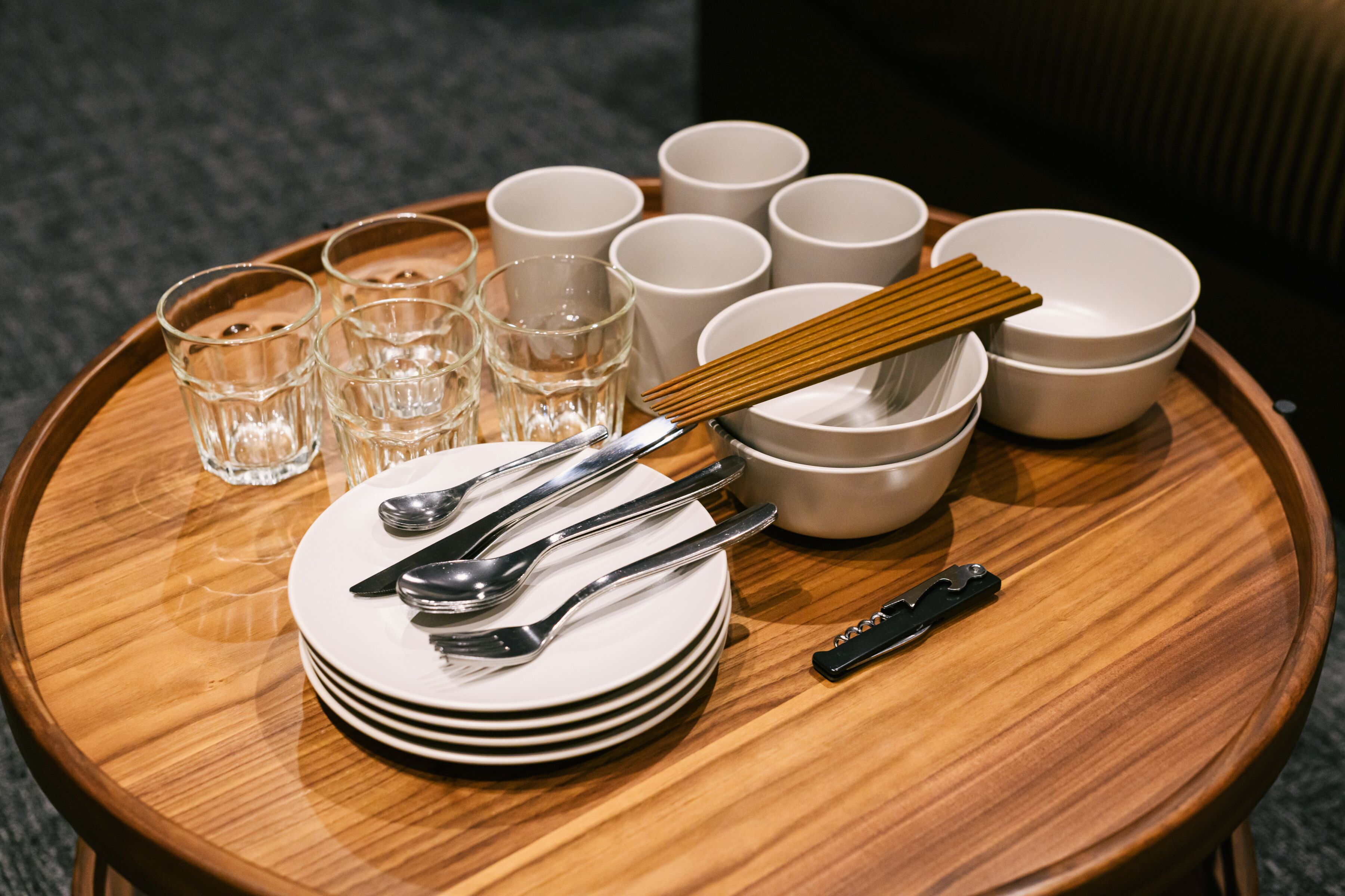 Une table ronde en bois, sur laquelle sont disposées des assiettes blanches empilées, des couverts en argent et des verres transparents.