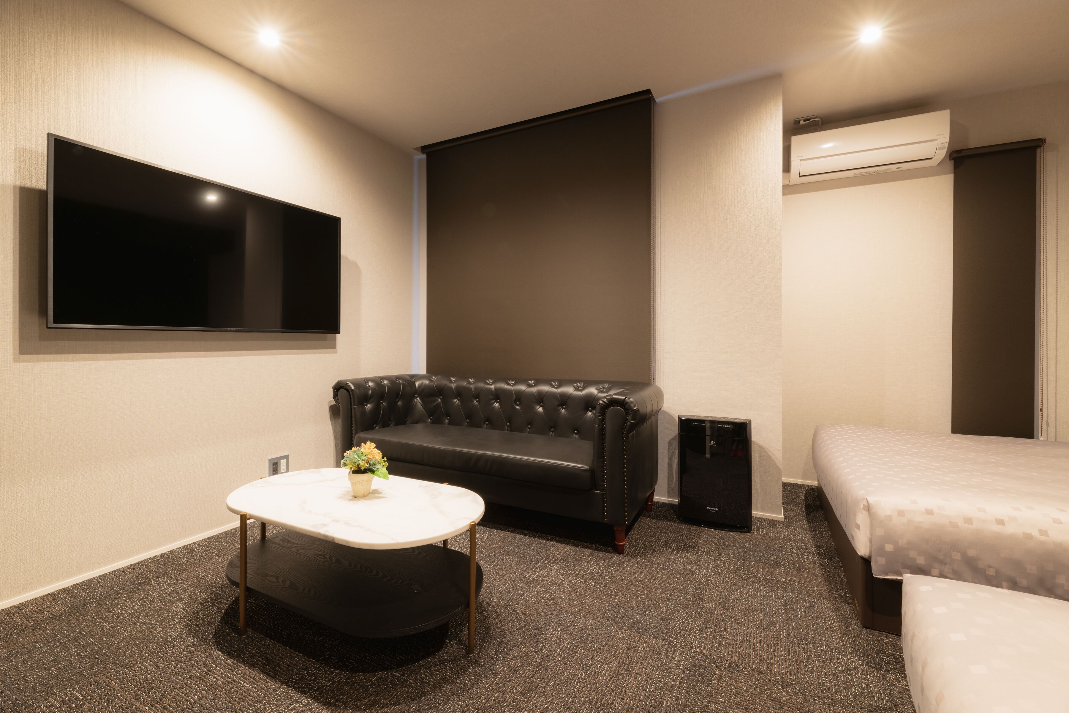 A modern hotel room with a flat-screen TV, a leather sofa, a round coffee table with flowers, a bed, and a carpeted floor.