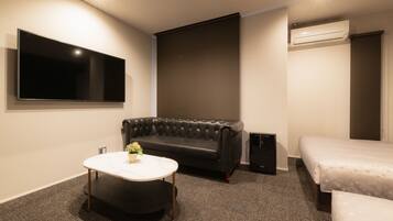 A modern hotel room with a flat-screen TV, a leather sofa, a round coffee table with flowers, a bed, and a carpeted floor.