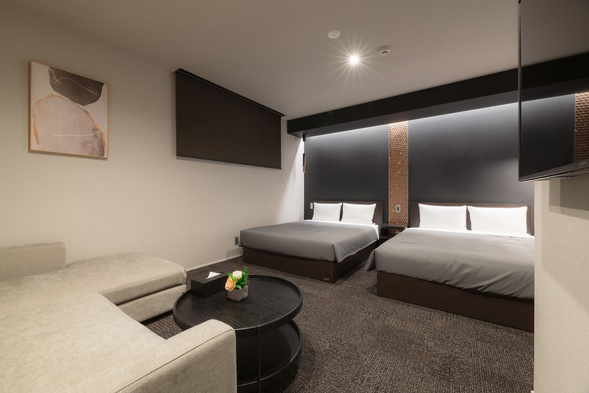 Une chambre d’hôtel moderne équipée de deux lits simples, d’un canapé, d’une table basse ronde et d’un téléviseur à écran plat fixé au mur.