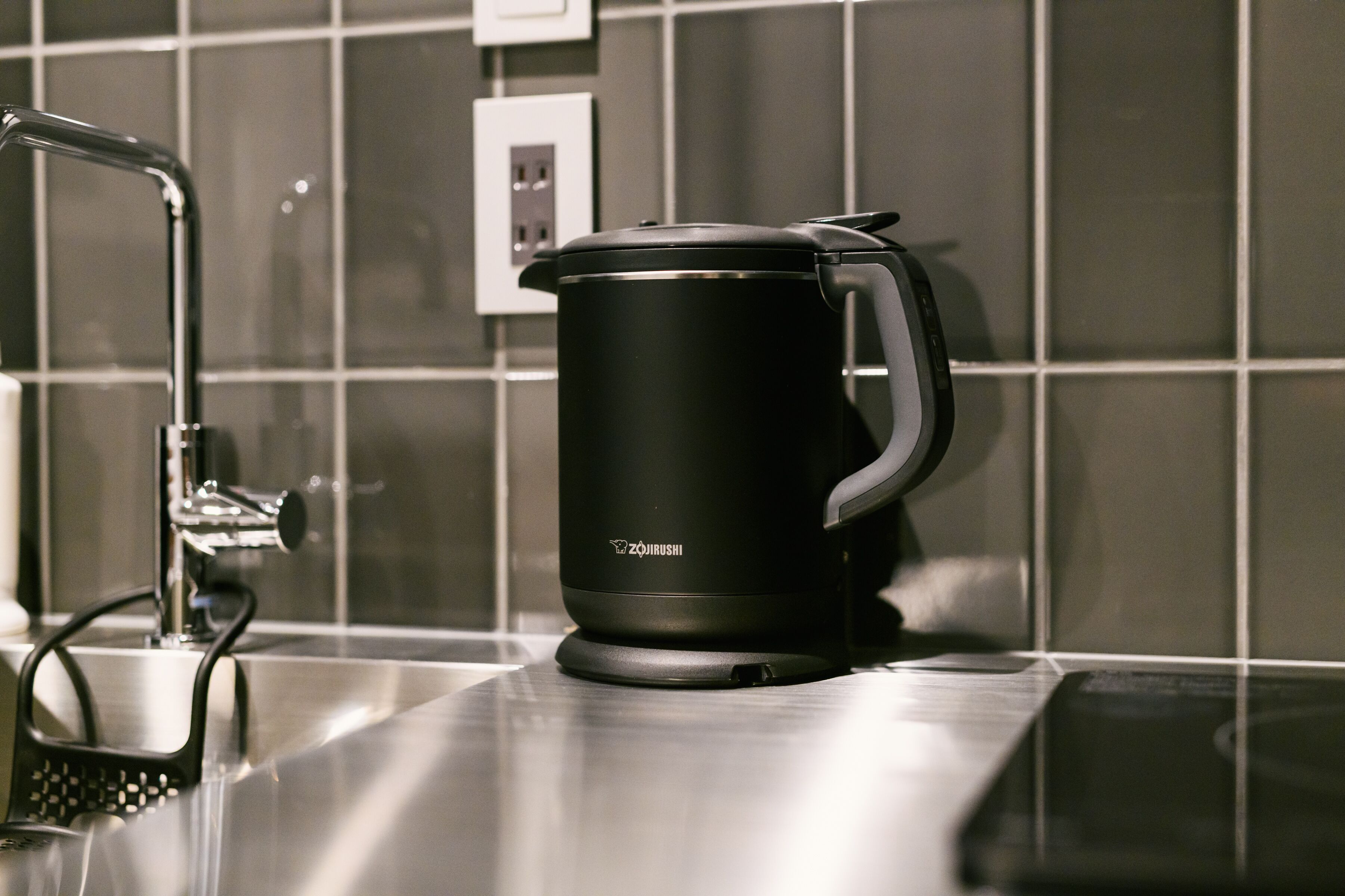 A black electric kettle on a stainless steel countertop with a modern faucet.