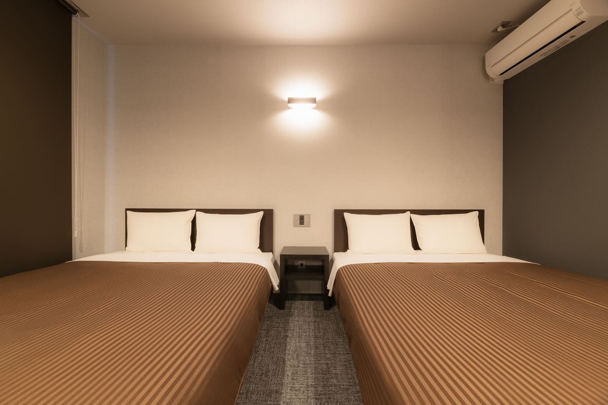 Une chambre d’hôtel avec deux lits, une table de chevet, une lampe fixée au mur et un appareil de climatisation.