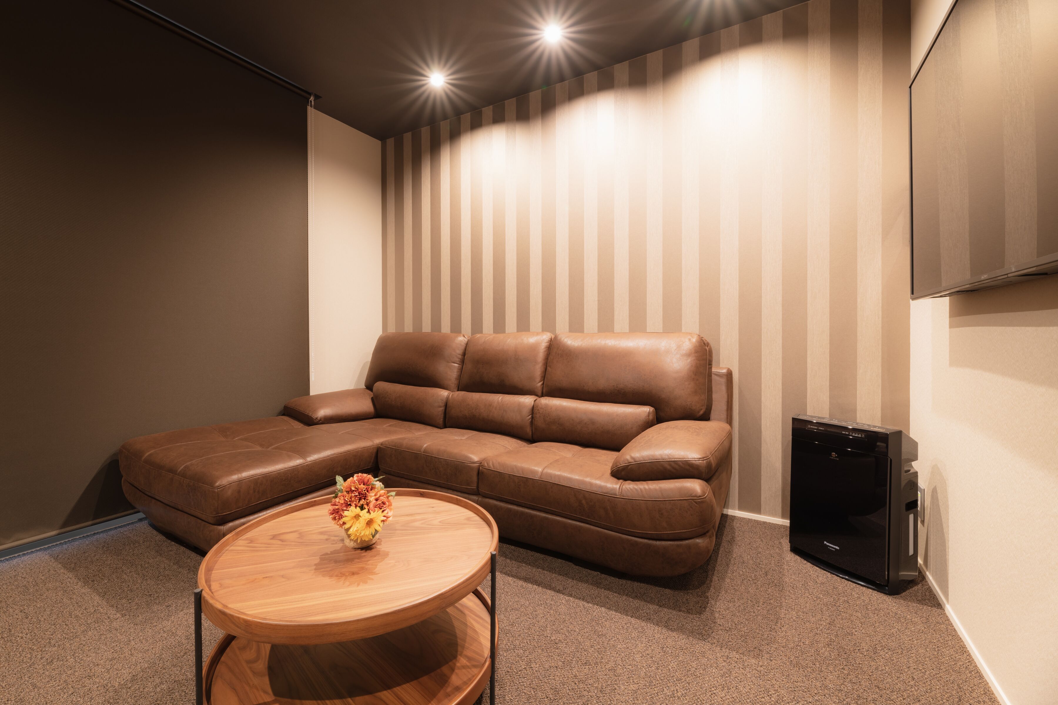 A modern living room with a brown leather sectional sofa, a wooden coffee table with a flower arrangement, and a flat-screen TV mounted on the wall.