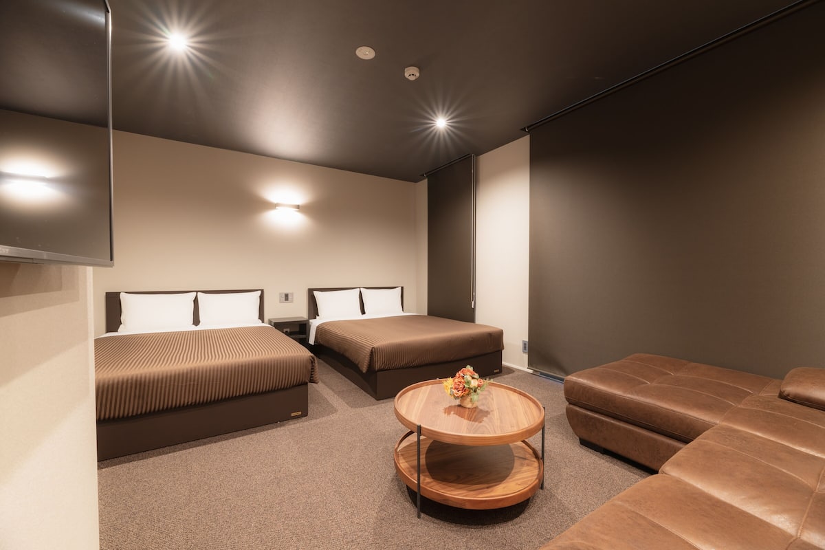 Une chambre d’hôtel moderne avec deux lits, un canapé, une table basse ronde en bois et un grand miroir.