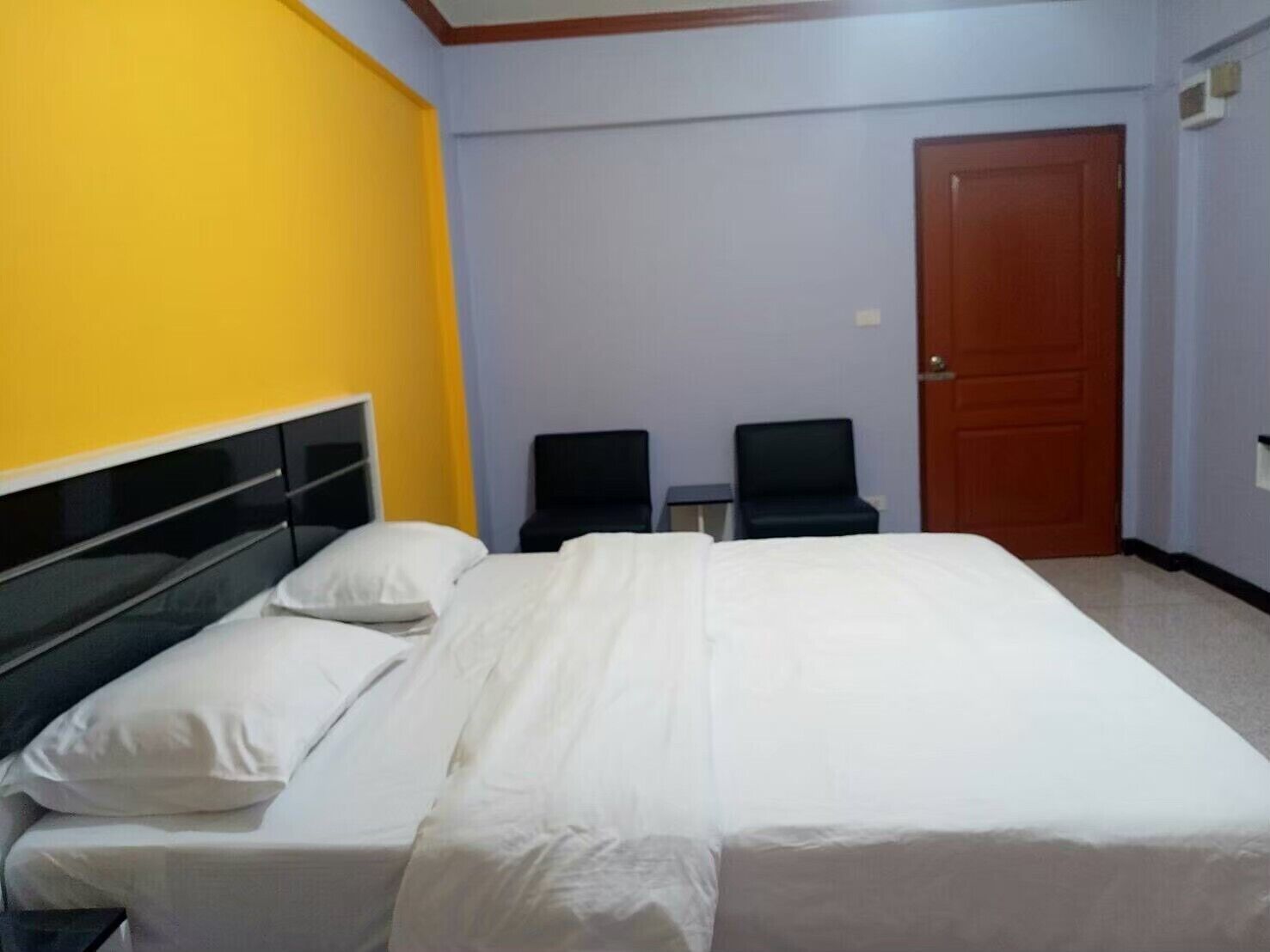 A hotel room with a bed, two chairs, a wooden door, and a yellow accent wall.