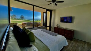 A bedroom with a large bed, a ceiling fan, a TV on the wall, and a view of a golf course through the window. - Gorgeous View- Beach Village at Palmas Del Mar (Palmas del Mar)