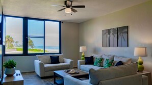 A modern living room with two sofas, a coffee table, and a ceiling fan. There are two lamps on side tables, and a large window with a view of the beach. - Gorgeous View- Beach Village at Palmas Del Mar (Palmas del Mar)