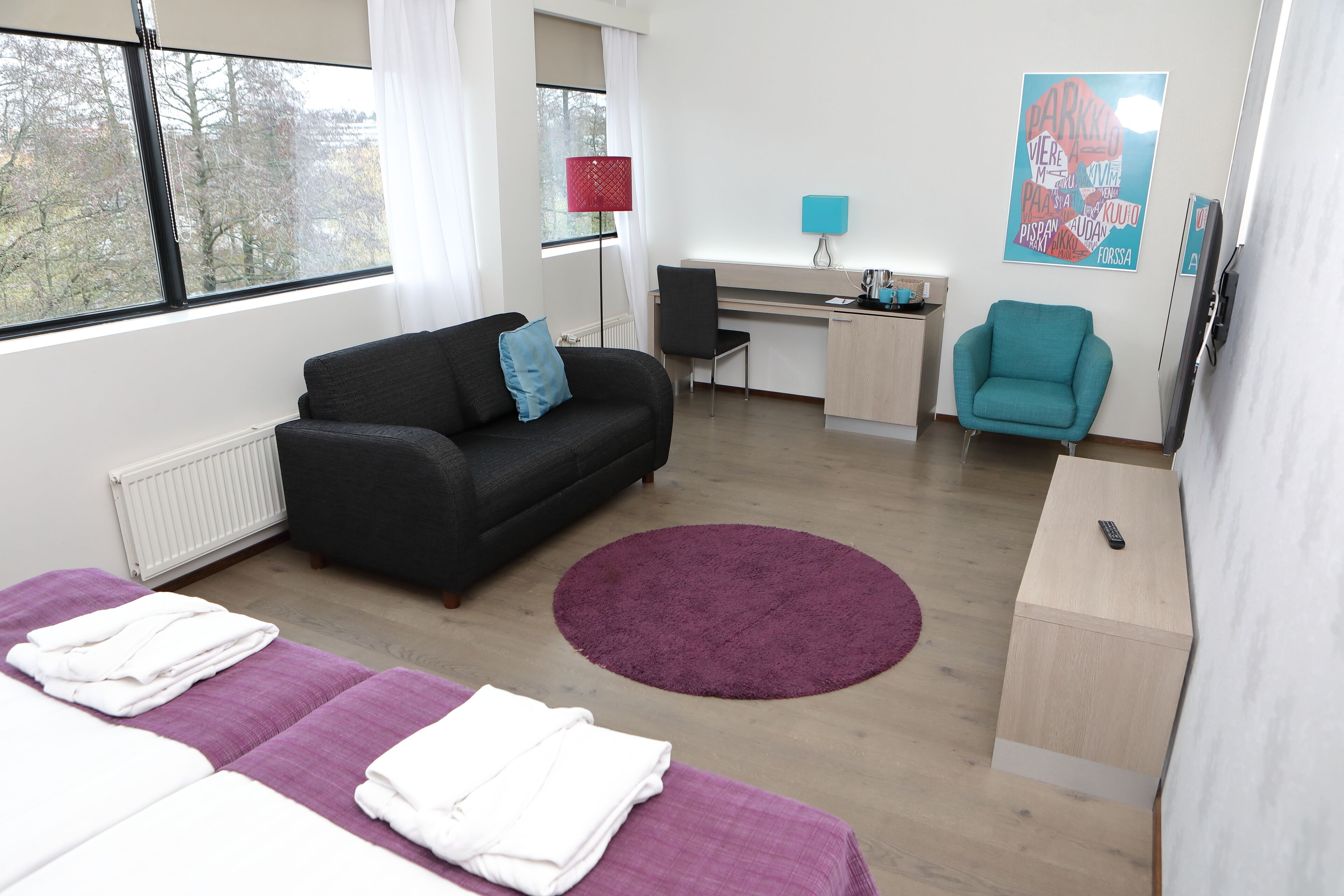 A modern hotel room with a black sofa, a teal armchair, a desk, and a bed with purple bedding.