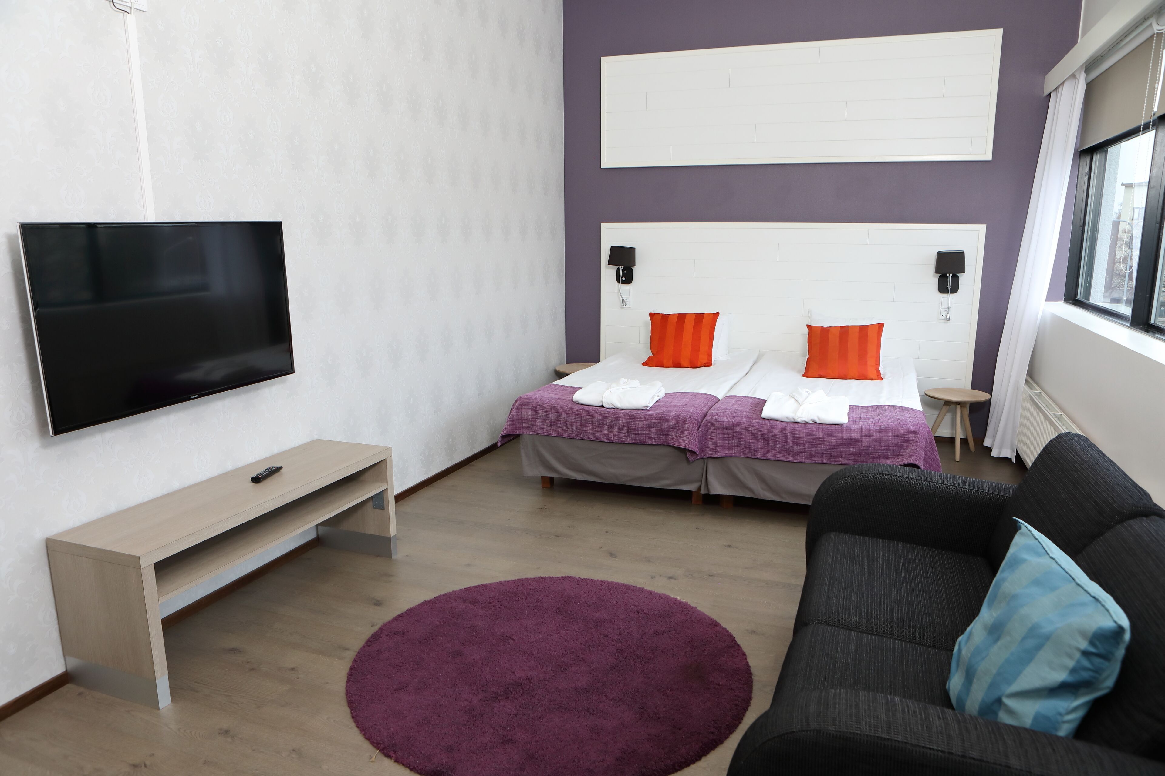 A modern hotel room with a large flat-screen TV, a bed with orange and white pillows, a dark sofa, and a purple rug.