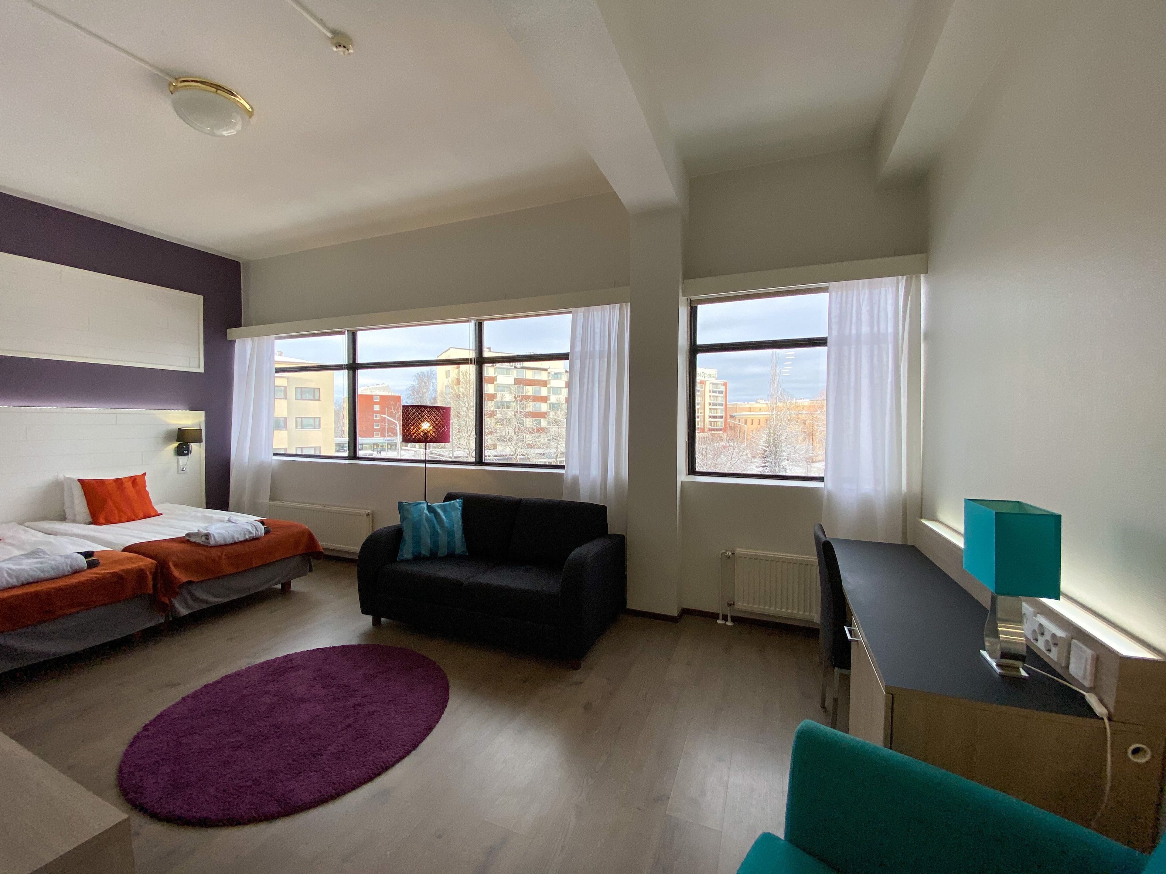 Habitación de hotel moderna con dos camas, un sofá, un escritorio y grandes ventanales con vistas a la ciudad.