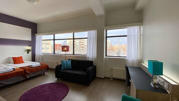 Habitación de hotel moderna con dos camas, un sofá, un escritorio y grandes ventanales con vistas a la ciudad.
